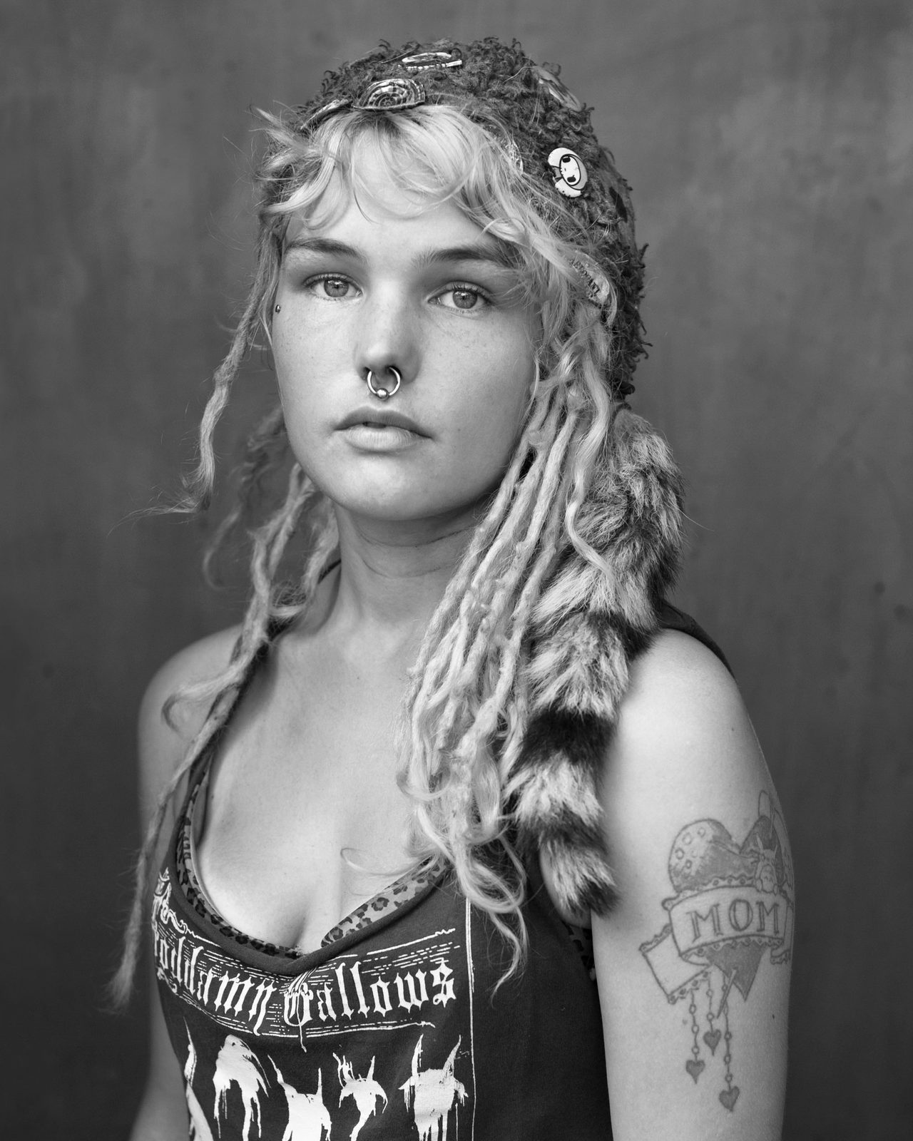 Black and white documentary portrait of a young female drifter looking directly into the camera, tattoos, nose piercing, dreadlocks and a handmade hat. RASKULL by Michael Joseph, Woman Portrait Gallery.
